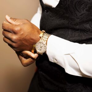 Man in elegant formal wear adjusts his gold watch while wearing a white shirt and black waistcoat. Perfect attire for a sophisticated and stylish event or business meeting.
