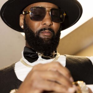 A stylish man with a beard wearing sunglasses a black hat and formal attire including a bow tie and gold jewelry displaying elegance and sophistication indoors.