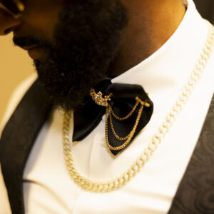 Close-up of a man wearing a stylish bow tie with layered gold chains and a patterned waistcoat over a white shirt showcasing elegant men's fashion accessories.