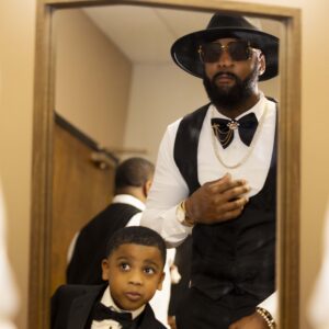 Father and son dressed in stylish suits pose confidently in front of a mirror. The father wears a hat and sunglasses, while the son smiles in a tuxedo.