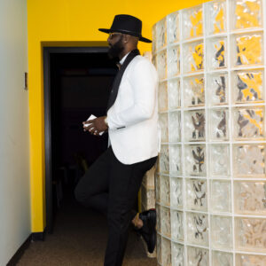 Man in stylish attire with black hat and white blazer leans against a decorative glass wall in a modern office space with a yellow accent wall.