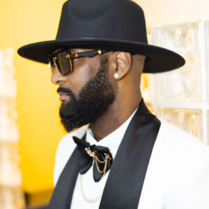 Stylish man with a beard wearing a black hat sunglasses white suit and black bow tie accessorized with gold and pearl jewelry set against a vibrant yellow background