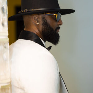 Stylish man in black fedora and sunglasses wearing a white suit and black bow tie standing indoors showcasing elegance and modern fashion trends.