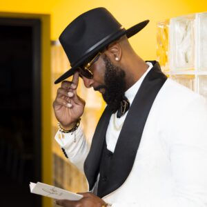Stylish man in a white suit with a black hat and sunglasses reading a paper in a brightly lit room. Fashionable attire blending elegance and modern style.