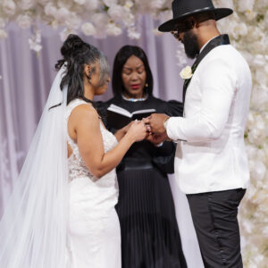 Bride and groom exchange vows in elegant wedding ceremony with floral backdrop officiated by a woman in black attire capturing a touching moment of love and commitment