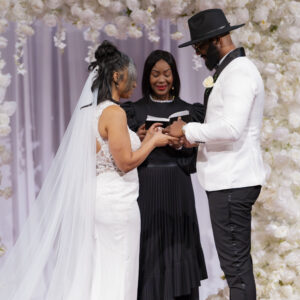 Bride and groom exchanging vows during a wedding ceremony with a floral arch background, officiant present, symbolizing love and commitment in an elegant setting.
