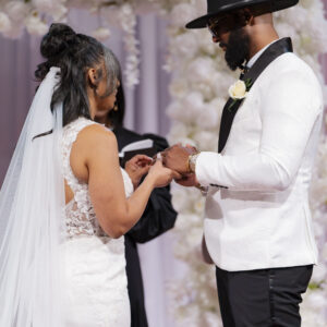 A couple exchanging rings during a wedding ceremony surrounded by elegant white floral decorations and draped curtains creating a romantic and intimate atmosphere.