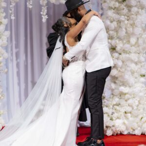 Bride and groom embrace in a romantic wedding scene on a flower-adorned stage. The bride wears a long white gown with a veil while the groom is dressed in a white jacket.