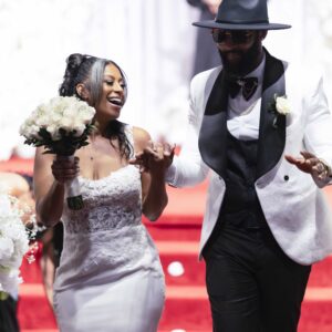 Happy couple celebrating their wedding day walking down a red carpet aisle bride holding a bouquet and groom in a stylish white jacket and black hat smiling joyfully