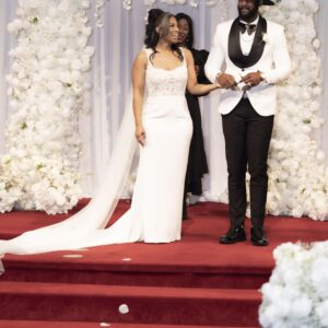 Bride and groom smiling and holding hands during their wedding ceremony standing on a red carpet with a beautiful floral backdrop covered in white flowers.