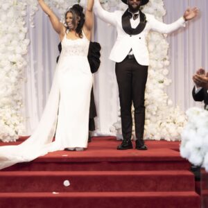 Bride and groom celebrate their wedding day on a red carpeted stage with white floral backdrop petals scattered on the floor capturing a joyful moment together