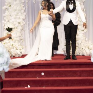 Bride and groom joyfully walking down the aisle on their wedding day surrounded by white floral decorations and red carpet setting for a romantic celebration.