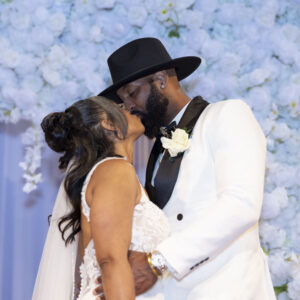 Bride and groom share a romantic kiss at a wedding ceremony with a backdrop of white florals. The groom is in a stylish black hat and white suit, and the bride in a lace gown.
