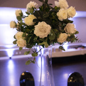 Elegant wedding reception table centerpiece featuring a tall vase with lush white roses and greenery enhancing the ambiance of a beautifully decorated event venue.