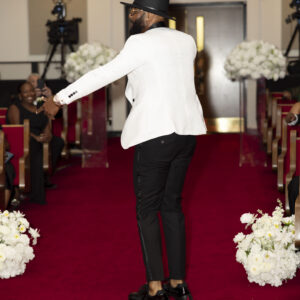 Man in a white blazer and black hat walking down a red-carpeted aisle surrounded by elegant white floral arrangements at a formal event venue with seated guests.