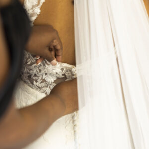 Bride getting dressed in a lace wedding gown with veil as someone helps fasten buttons adding final touches before walking down the aisle on her special day