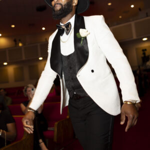 Stylish man in a white tuxedo with a black lapel and bow tie wearing sunglasses and a hat walking confidently in a formal setting with red carpeted seating.