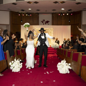 Bride and groom joyfully exit ceremony hall surrounded by cheering guests and flower petals on red carpet creating a festive atmosphere on their wedding day.