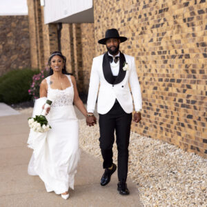 Elegant bride and groom walking hand in hand outside a brick building on their wedding day. Bride in a white dress with bouquet and groom in a stylish white tuxedo.