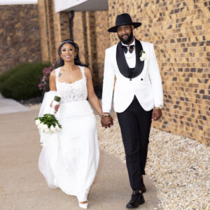 Elegant bride and groom walking hand in hand on their wedding day. Bride wears a white gown holding a bouquet. Groom dressed in a white tuxedo with a black hat.