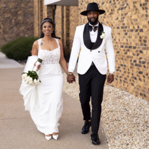 Bride and groom walking hand in hand on their wedding day bride in white gown with flowers and groom in a stylish white jacket and black hat against brick wall backdrop