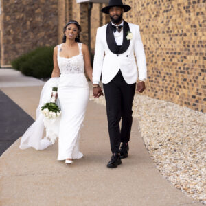 Elegant couple in wedding attire walking hand in hand on a pathway including a bride in a white gown and veil and groom in a white tuxedo and black hat holding a bouquet