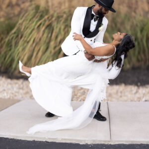 A joyful bride and groom share a romantic dance move outdoors, with the groom effortlessly dipping the bride in her elegant white gown, both smiling and happy.