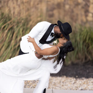 Groom in white suit and black hat romantically dips bride in white wedding dress for kiss in scenic outdoor setting with blurred natural background.
