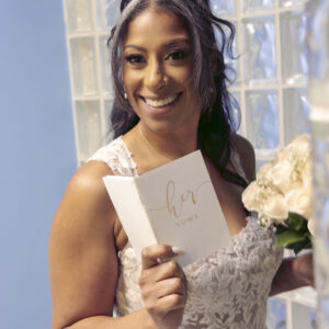 Smiling bride in a lace wedding dress holding her vows and a bouquet of white flowers in a bright room with a glass block wall.