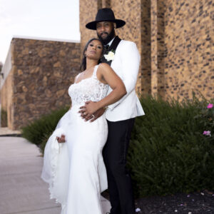 Bride in elegant white gown and groom in stylish black hat and suit, posing happily outdoors beside a textured stone building, capturing their special wedding day.