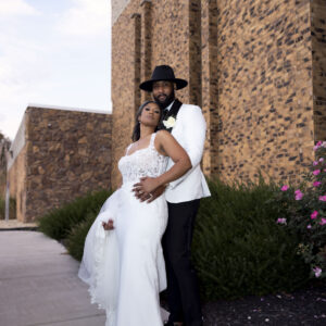 A couple stands in elegant wedding attire near a modern brick building with blooming flowers. The bride wears a white gown and the groom is in a stylish suit and hat.