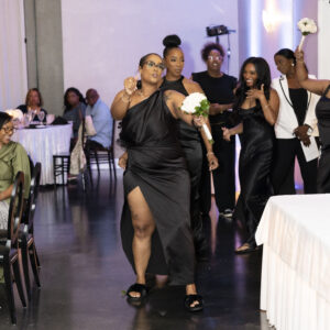 Group of people in elegant black attire walk confidently through a modern event space, with one holding a bouquet, creating a lively and joyful atmosphere.