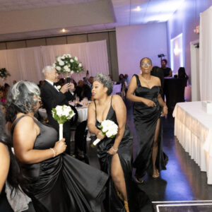 Women in elegant black dresses dance joyfully holding white flowers at a formal event with a sophisticated ambiance and seated guests in a beautifully decorated venue.