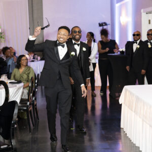 A joyful group of men in formal wear celebrating at a wedding reception indoors in a beautifully decorated venue with guests clapping and smiling in the background.