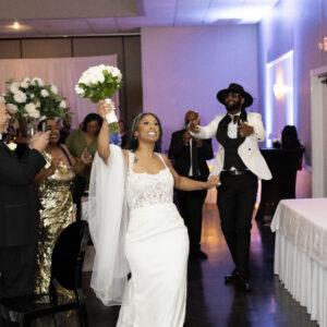 Bride joyfully entering wedding reception holding bouquet followed by a jubilant groom in black and white suit while guests celebrate in elegantly decorated venue.