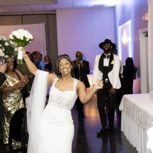Bride celebrating at a wedding reception with guests applauding and cheering in an elegant venue featuring modern white decor and ambient lighting.