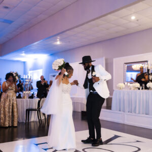 Bride and groom joyfully dance at a wedding reception with guests cheering, elegant decor, and white floral arrangements creating a festive and romantic atmosphere.