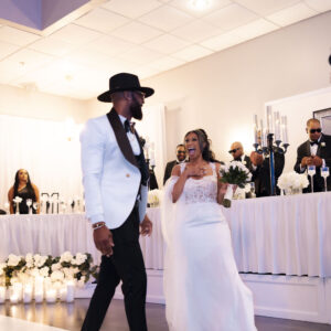 Elegant couple dancing at a wedding reception dressed in stylish formal attire with a smiling bridal party in the background amid a beautifully decorated venue.