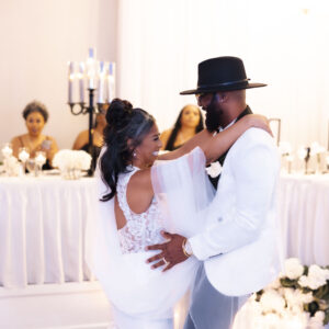 Newlywed couple sharing a romantic dance during wedding reception with elegantly dressed guests seated at a beautifully decorated table in the background.