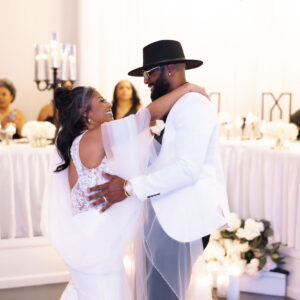 Bride and groom share a joyful dance at their wedding reception in an elegant venue with guests watching. The couple is dressed in stylish white attire.
