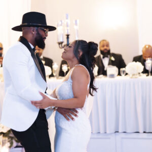 Newlywed couple shares a joyful first dance at a wedding reception with elegantly decorated venue and smiling guests seated in the background wearing formal attire.
