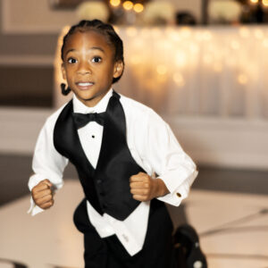 Young boy in a black tuxedo vest and bow tie joyfully running on a decorated dance floor at a celebratory event showcasing elegance and festive atmosphere.
