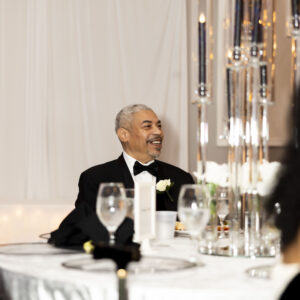 Smiling man in a tuxedo seated at an elegant table setting with candles and glassware reflecting a warm ambiance at a formal event or celebration.