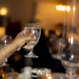 A raised hand holding a glass of water in a dimly lit elegant dining setting with glasses and candles on the table, creating a warm and festive atmosphere.