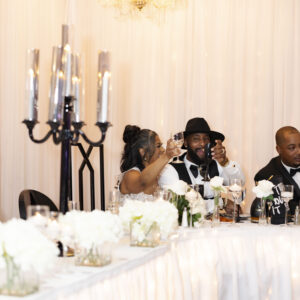 People celebrating at a beautifully decorated wedding reception with elegant table settings and floral arrangements, raising glasses in a toast under warm lighting.