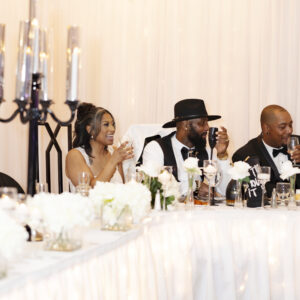 Guests at a wedding reception sit at a beautifully decorated table laughing and enjoying drinks in an elegant venue with white floral arrangements and candlelit ambiance.
