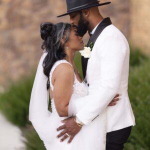 Bride and groom embrace lovingly in an elegant setting showcasing wedding attire with the groom in a stylish hat and the bride in a beautiful lace gown and veil.