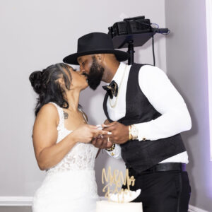 Bride and groom share a romantic kiss at their wedding reception standing next to a beautifully decorated cake symbolizing love and union on their special day.