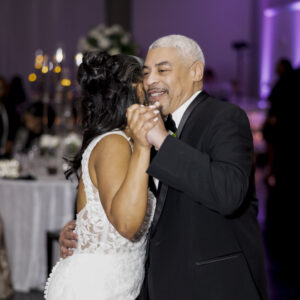 A bride in a white lace dress dances with an older man in a suit at a beautifully decorated wedding reception. They are smiling under soft purple lighting.