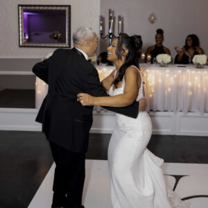 Bride in a white gown joyfully dances with an older man in a suit at a wedding reception. Guests are seated at a decorated table, softly lit with string lights.